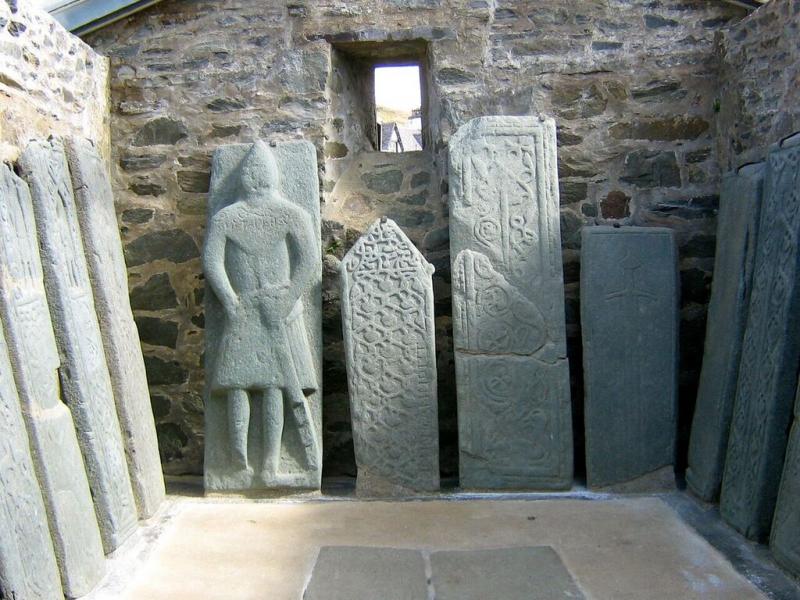 Ten graveslabs, including one with a carved knight effigy, propped up vertically, leaning against three walls of the interior of a building with a glass roof